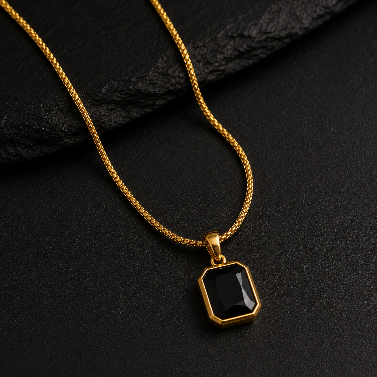 A gold chain necklace with a faceted black gemstone pendant, displayed on a dark textured slate surface.