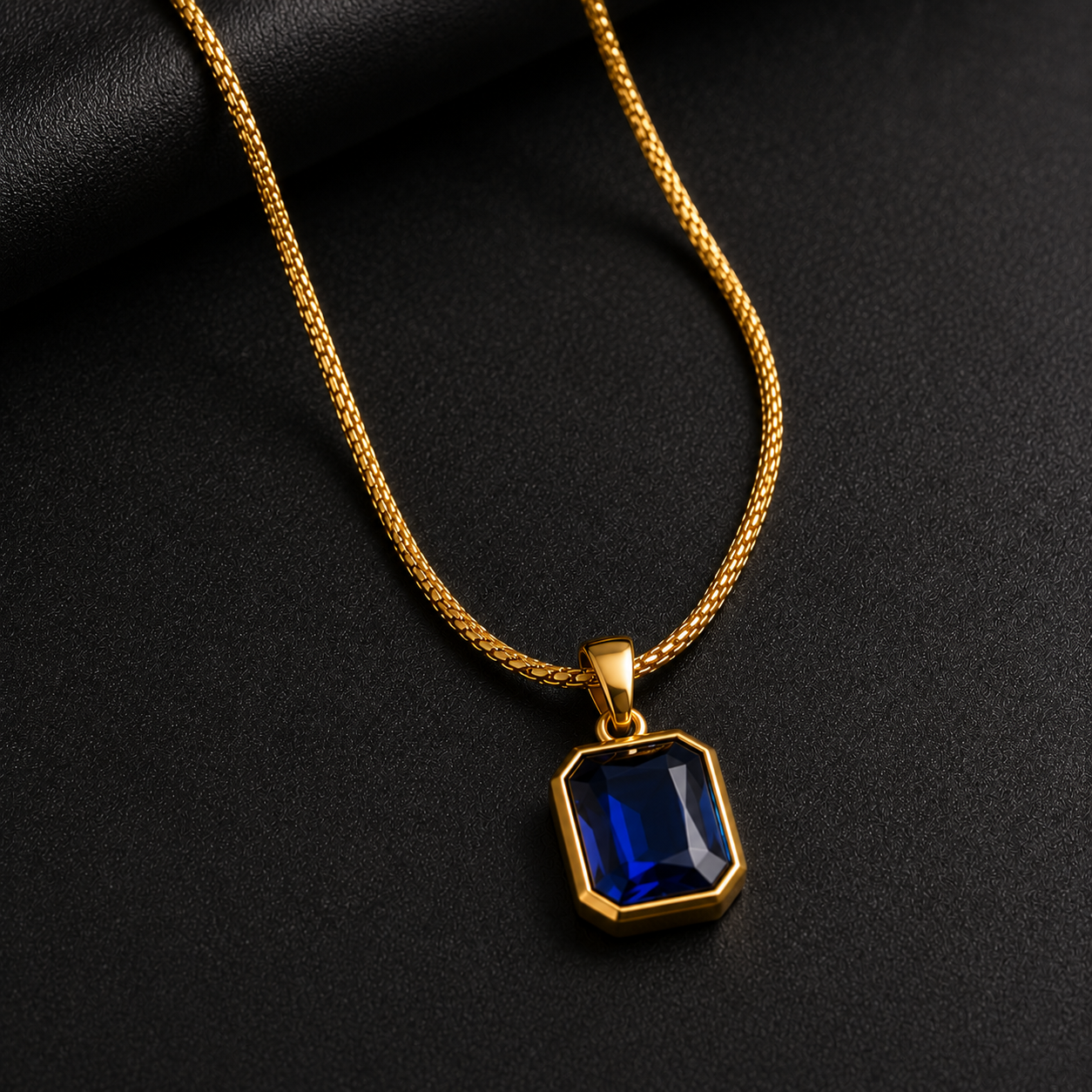A gold chain necklace with a rectangular gold pendant featuring a faceted deep blue gemstone, displayed on a textured black background.