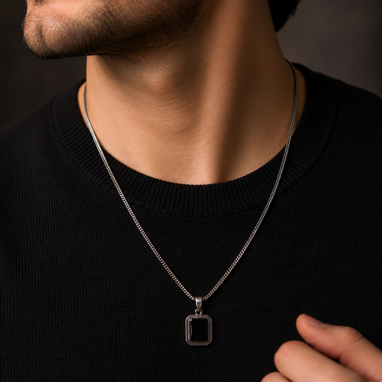 A close-up of a man wearing a silver chain necklace with a rectangular black pendant, styled with a black crewneck sweater.