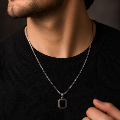 A close-up of a man wearing a silver chain necklace with a rectangular black pendant, styled with a black crewneck sweater.