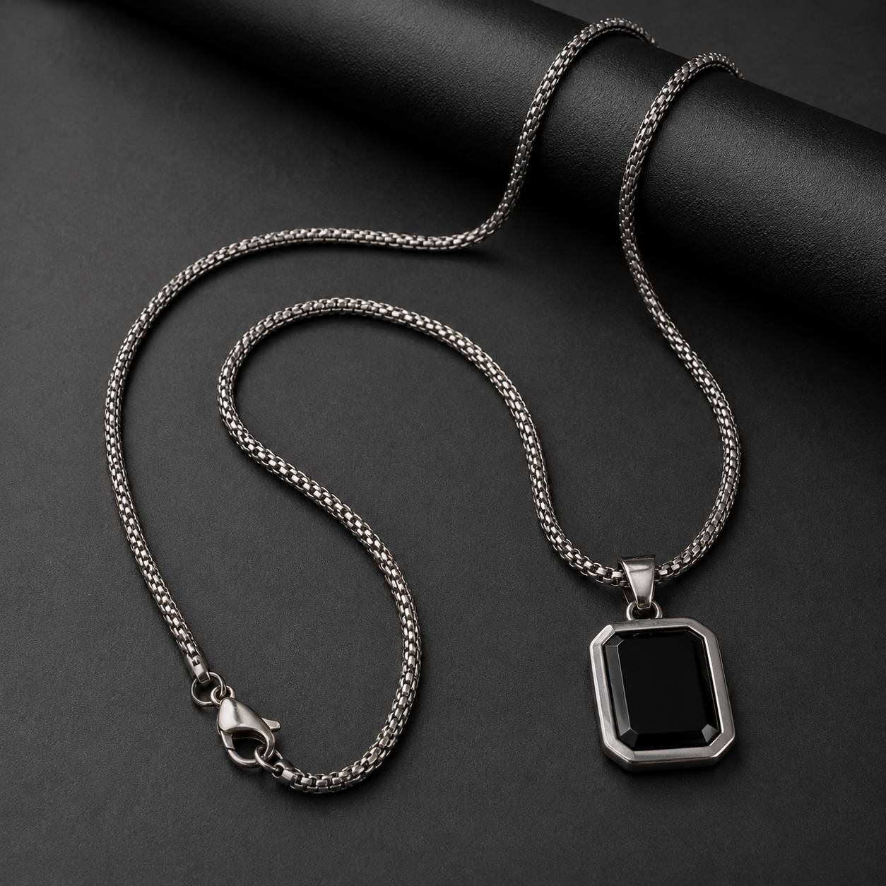 A silver-toned chain necklace with a geometric black pendant, displayed on a dark textured background.