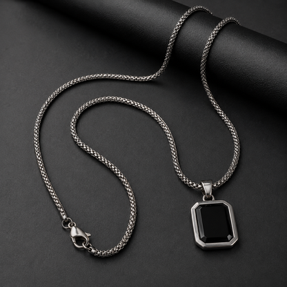 A silver-toned chain necklace with a geometric black pendant, displayed on a dark textured background.
