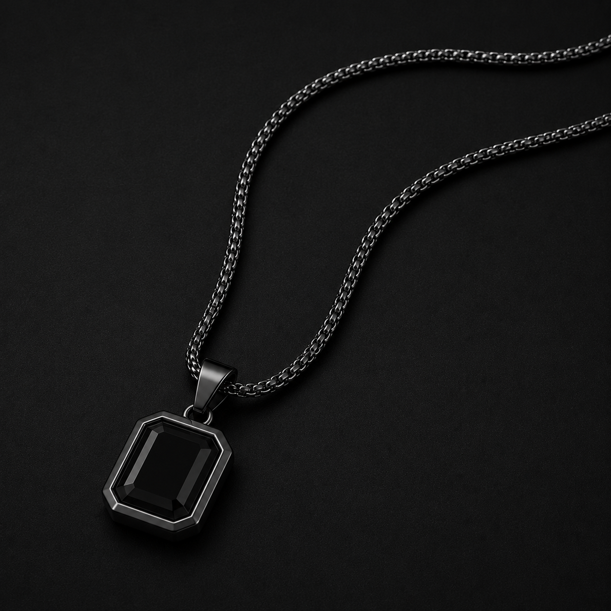 A men's pendant necklace featuring a rectangular faceted black gemstone in a dark gunmetal silver bezel, suspended from a matching dark silver chain against a solid black background.