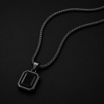 A men's pendant necklace featuring a rectangular faceted black gemstone in a dark gunmetal silver bezel, suspended from a matching dark silver chain against a solid black background.