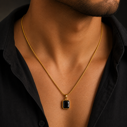 A close-up of a man's neck and chest wearing a gold chain necklace with a rectangular black pendant on a dark shirt.
