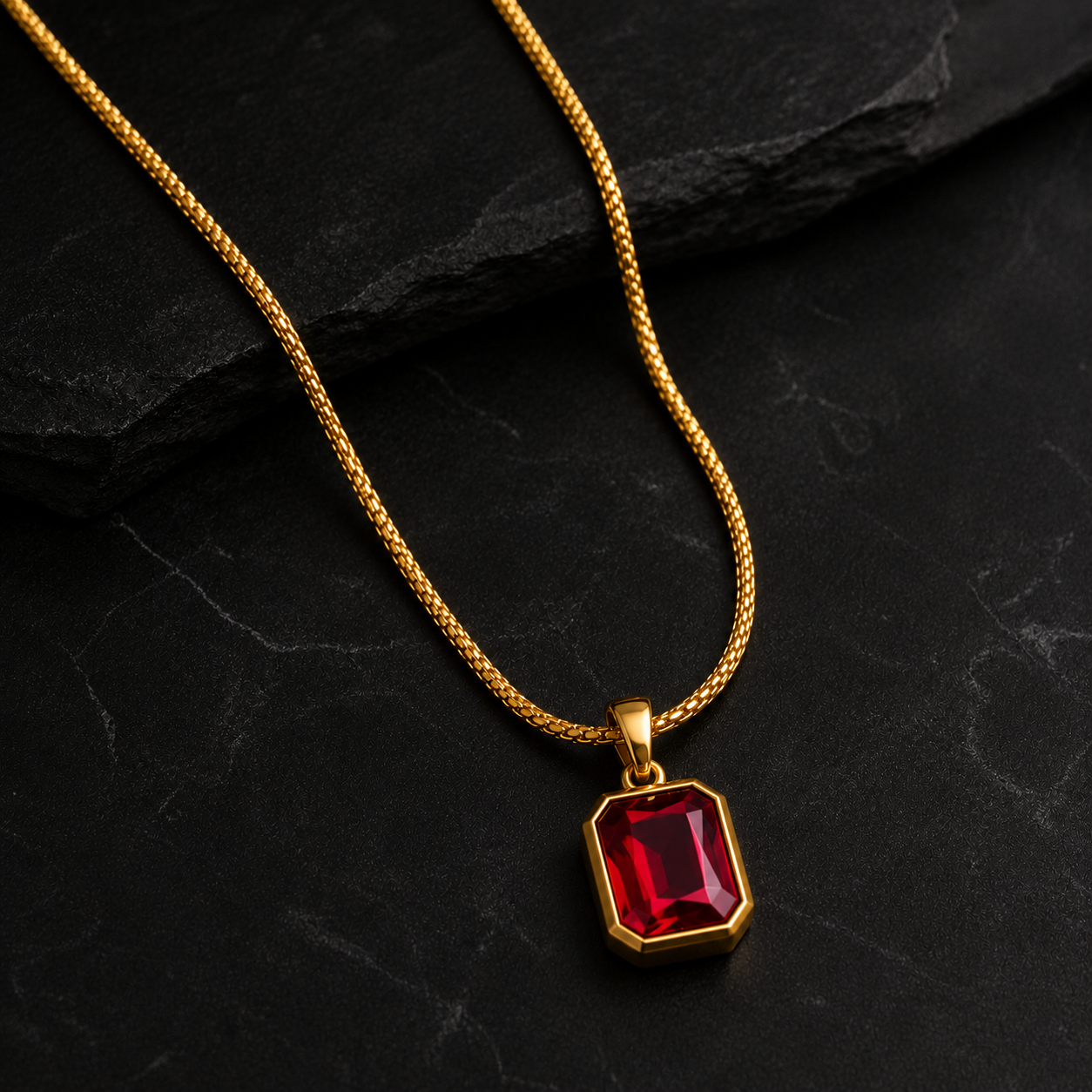 A gold chain necklace with a faceted red gemstone pendant, displayed on a dark textured slate background.