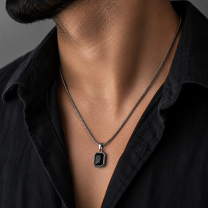A close-up of a man's neck and chest wearing a silver-toned chain necklace with a rectangular black pendant, paired with a black collared shirt.