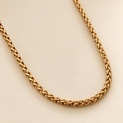 A close-up of a gold-toned braided chain necklace, likely a Cuban link style, arranged in a U-shape against a solid, warm off-white background.